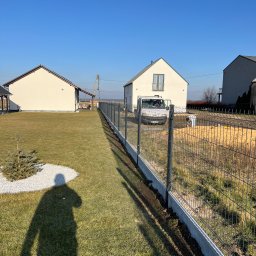 PanelExpress Ogrodzenia - Nowoczesne ogrodzenie panelowe z podmurówką betonową na trawniku, w tle dom jednorodzinny i błękitne niebo. Widoczny cień osoby.