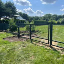 Nowoczesna, grafitowa brama panelowa z furtką, widoczna na tle zielonej trawy i drzew. Element ogrodzenia panelowego z siatki, brama otwarta, słoneczny dzień.