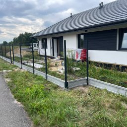 PanelExpress Ogrodzenia - Wznoszenie ogrodzenia panelowego z podmurówką prefabrykowaną przy posesji. Widoczne słupki i częściowo zamontowane panele na tle domu i drogi.