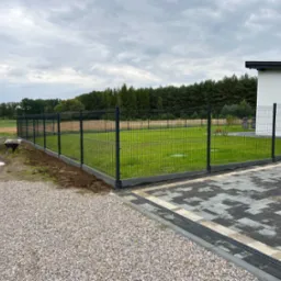 Nowoczesne ogrodzenie panelowe w kolorze grafitowym, oddzielające posesję z trawnikiem od ścieżki z kamienia. Widoczny fragment budynku i samoch&oacute;d dostawczy.