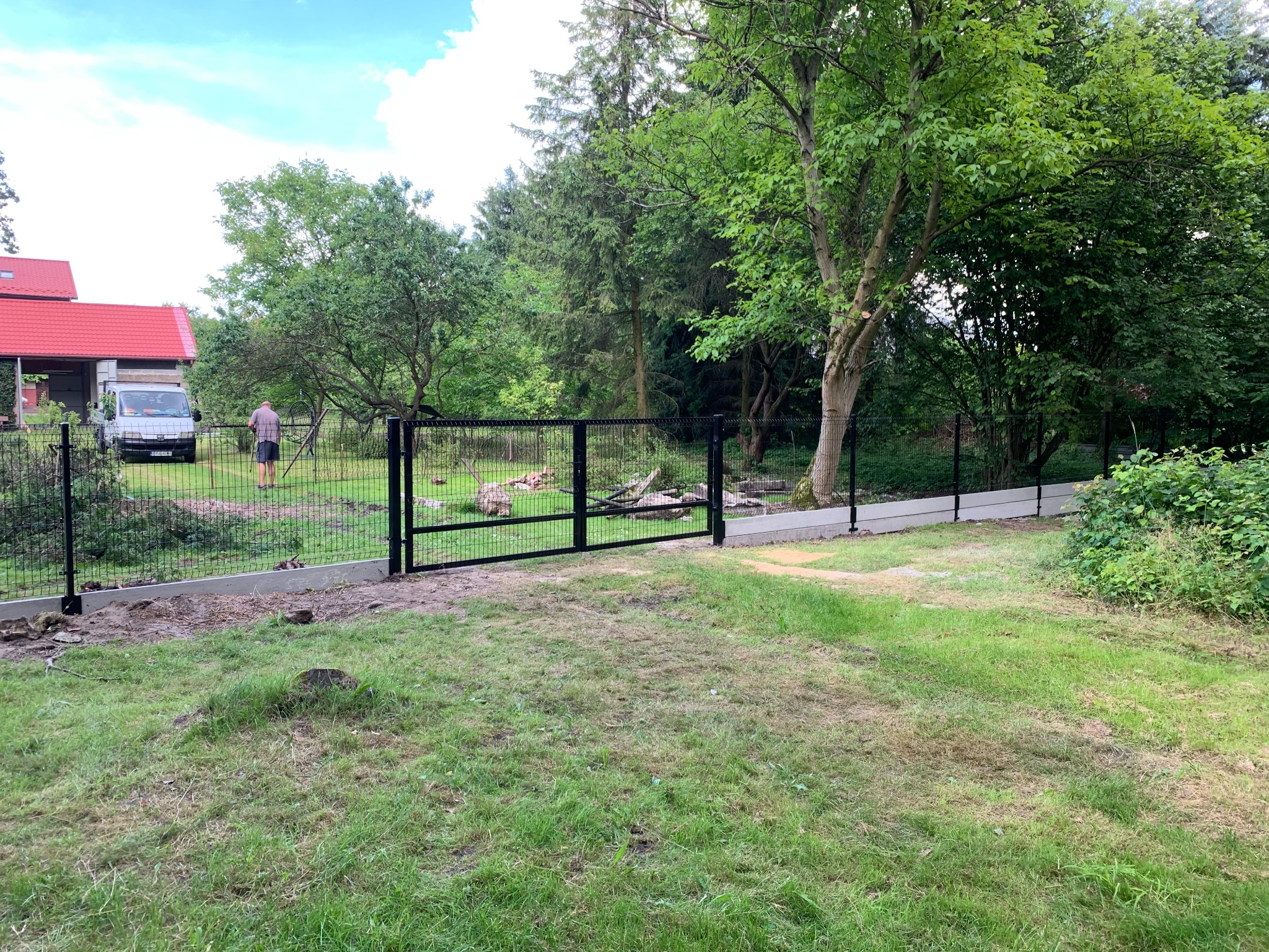 Czarna brama panelowa z siatki na betonowej podmurówce, widok na posesję z zielonym trawnikiem, drzewami i zaparkowanym białym dostawczakiem w tle, podczas montażu przez pracownika.