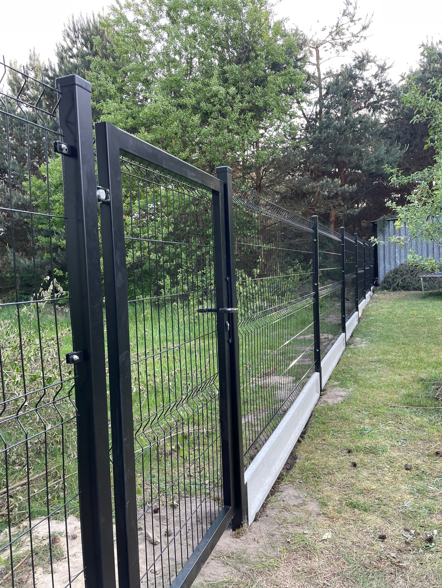 Nowoczesne, czarne ogrodzenie panelowe z otwartą furtką, osadzone na betonowej podmurówce, widoczne na tle zielonego trawnika i drzew.