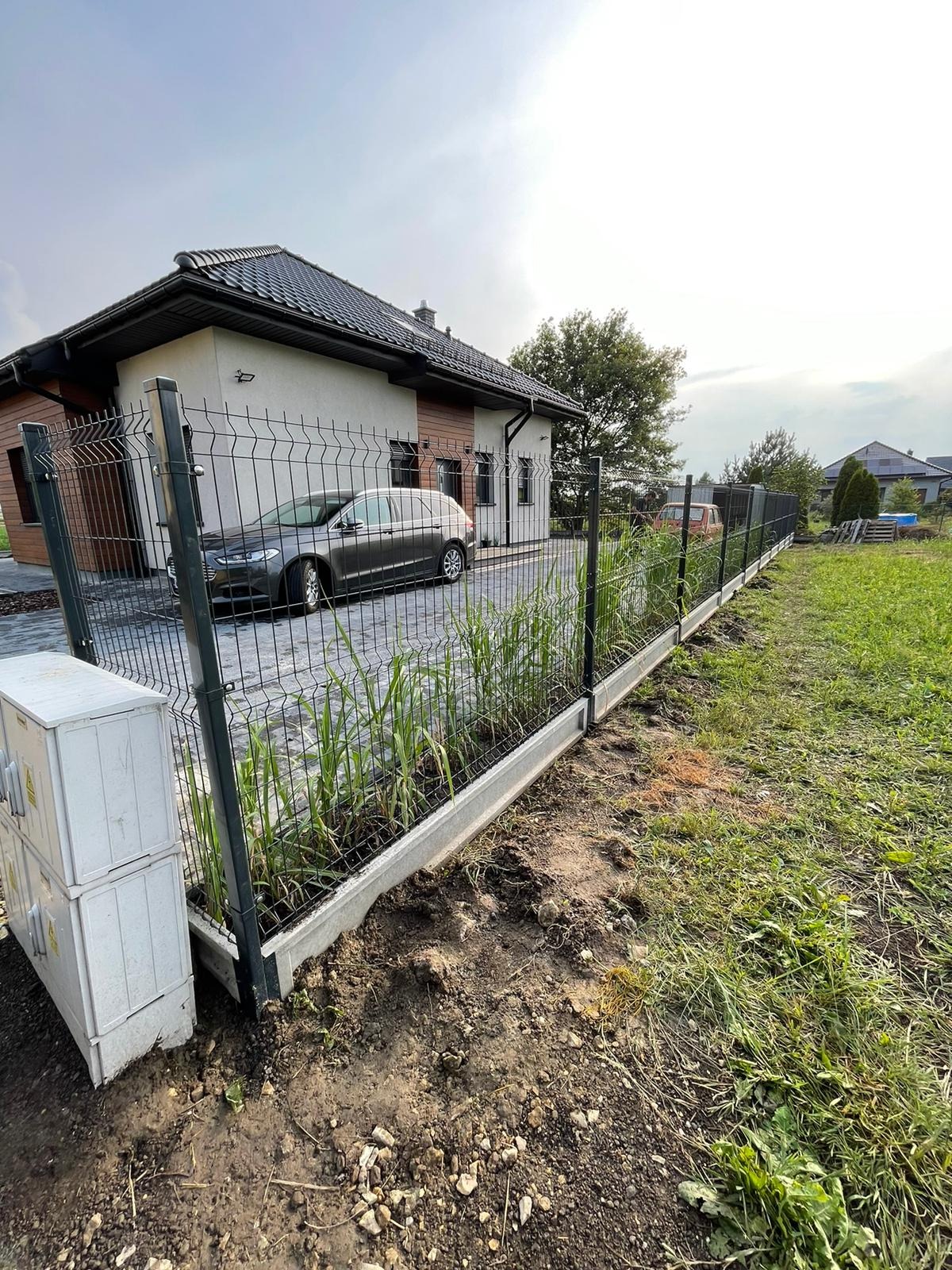 Nowo postawione ogrodzenie panelowe z podmurówką betonową, widoczne trawy rosnące wzdłuż ogrodzenia, dom jednorodzinny w tle, częściowo podjazd z zaparkowanym samochodem.