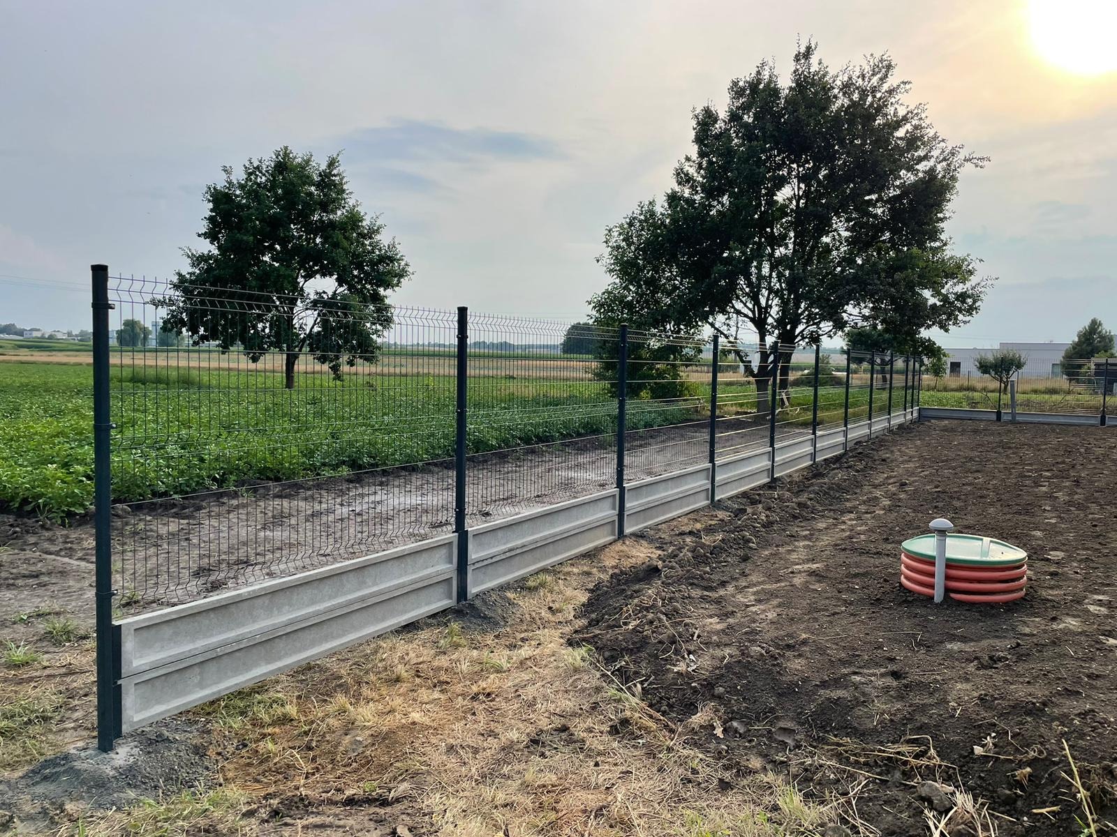 Nowo postawione ogrodzenie panelowe z podmurówką betonową na skraju pola uprawnego i działki z widocznym zbiornikiem na deszczówkę.