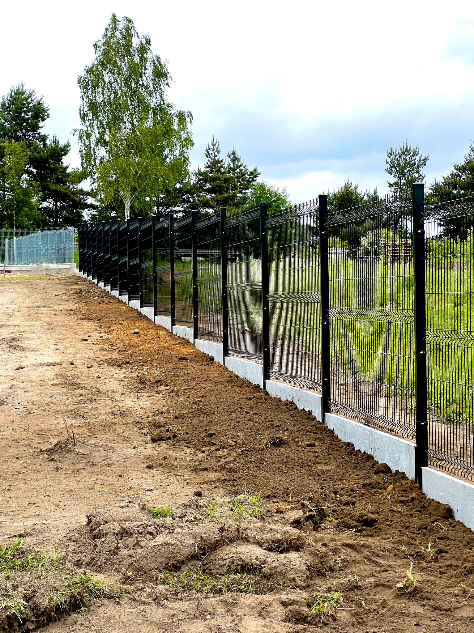 Nowo zamontowane, czarne ogrodzenie panelowe z podmurówką betonową na tle zieleni. Widok z perspektywy gruntu, podkreślający solidność konstrukcji.