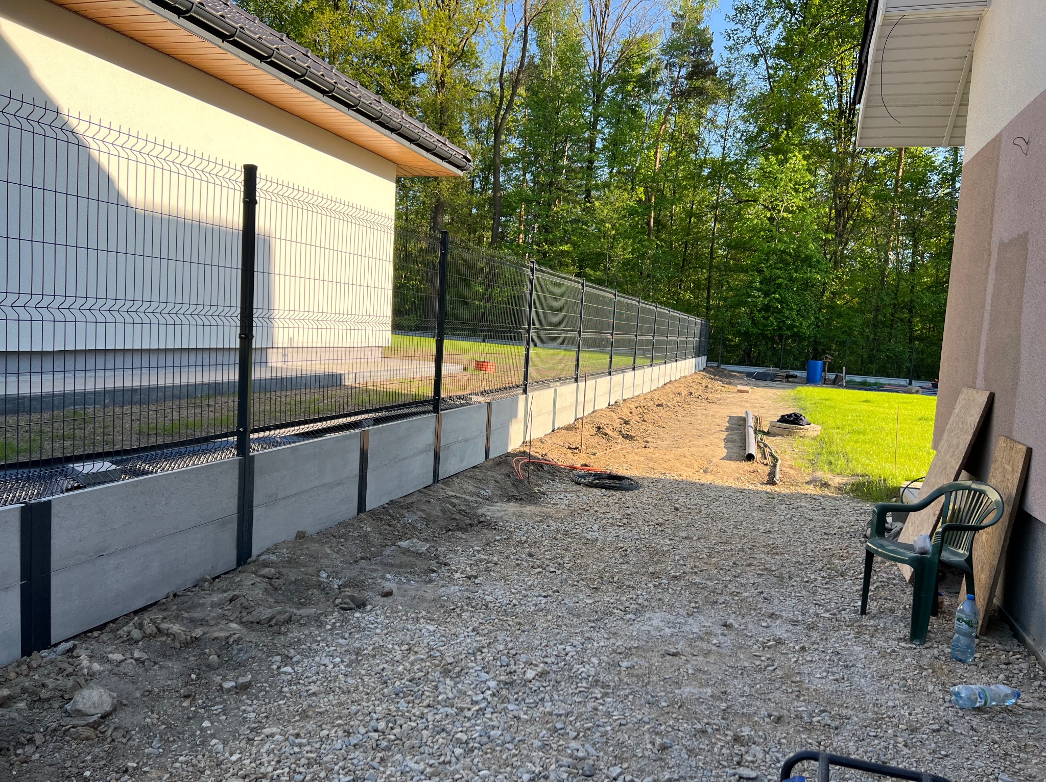 Nowoczesne ogrodzenie panelowe z podmurówką betonową, widok z boku na posesji. Ziemia wysypana żwirem, w tle zieleń drzew.