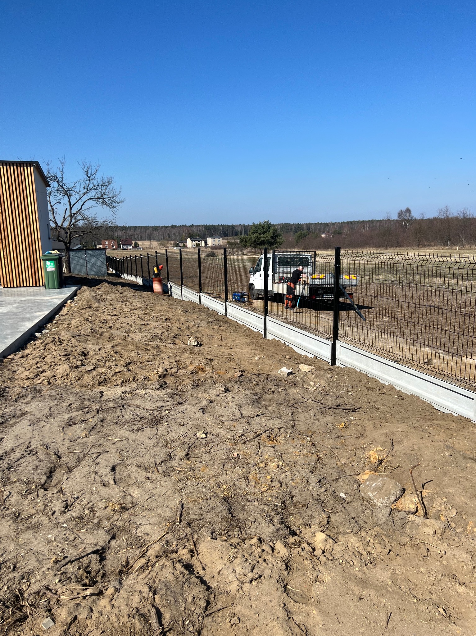 Montaż ogrodzenia panelowego na betonowej podmurówce. Pracownik przy ciężarówce. Widok na otwarty teren i budynek z drewnianymi elementami.