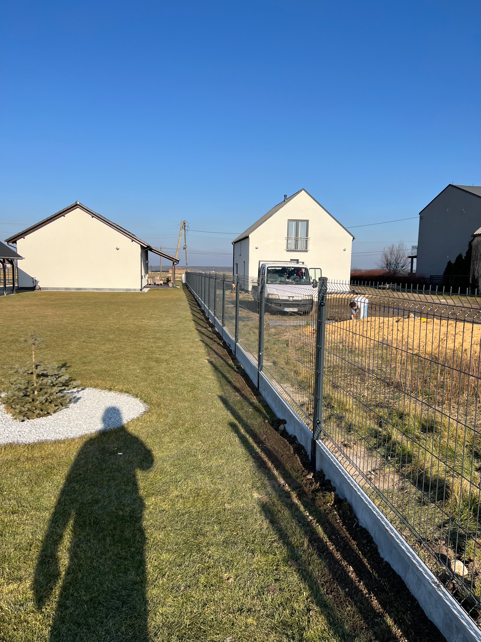 Nowoczesne ogrodzenie panelowe z podmurówką betonową na trawniku, w tle dom jednorodzinny i błękitne niebo. Widoczny cień osoby.