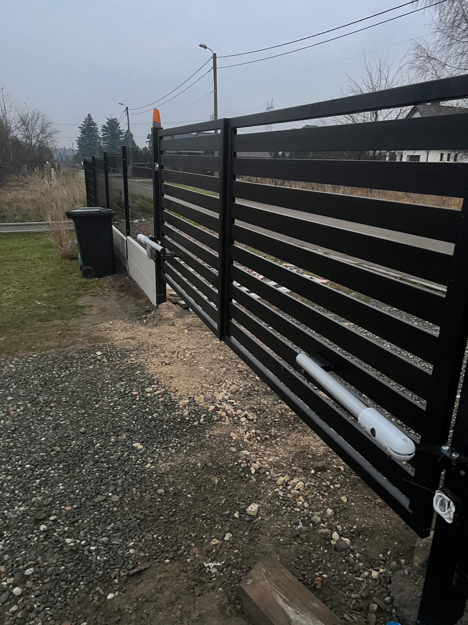 Nowoczesna, czarna brama wjazdowa z automatyką, widoczna z bliska na tle szarego nieba i otoczenia w trakcie montażu, z widocznym mechanizmem otwierania.