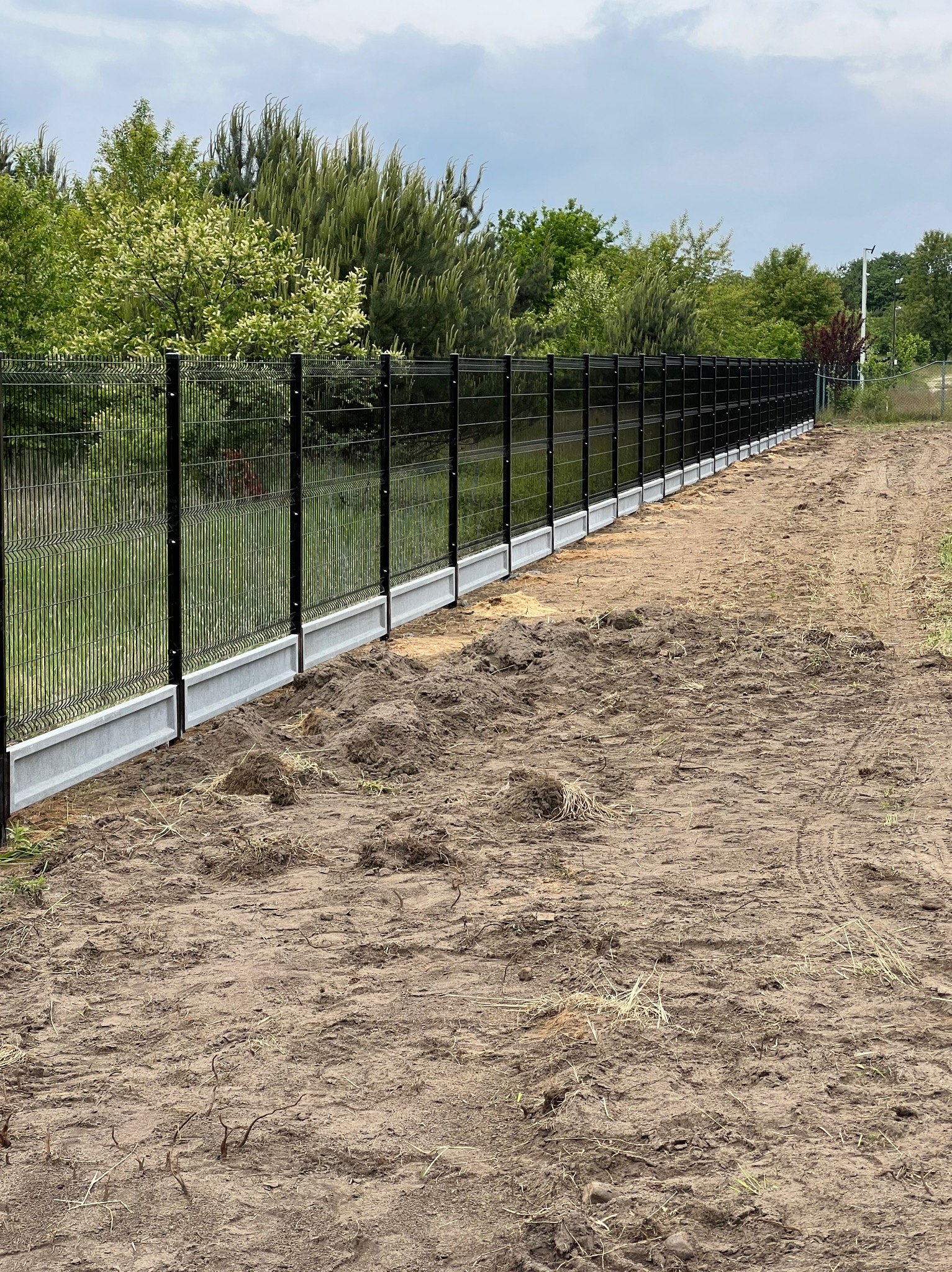 Nowoczesne ogrodzenie panelowe z podmurówką betonową, widok z boku na tle zieleni. Teren wokół świeżo wyrównany, przygotowany pod zagospodarowanie.