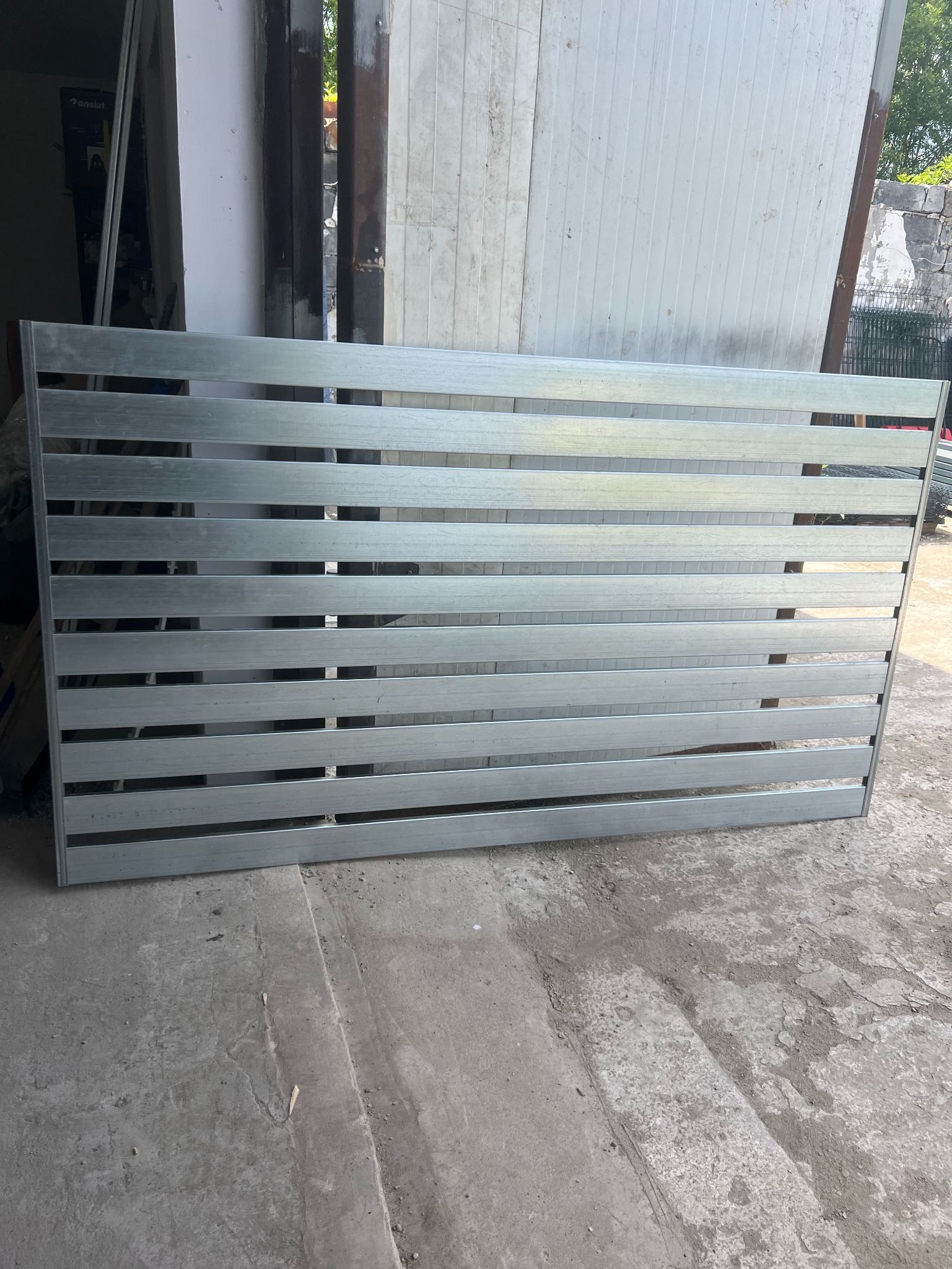 Szara, metalowa brama wjazdowa z poziomymi szczeblami, leżąca na betonowym podłożu w warsztacie, gotowa do montażu. Widoczne elementy konstrukcyjne.