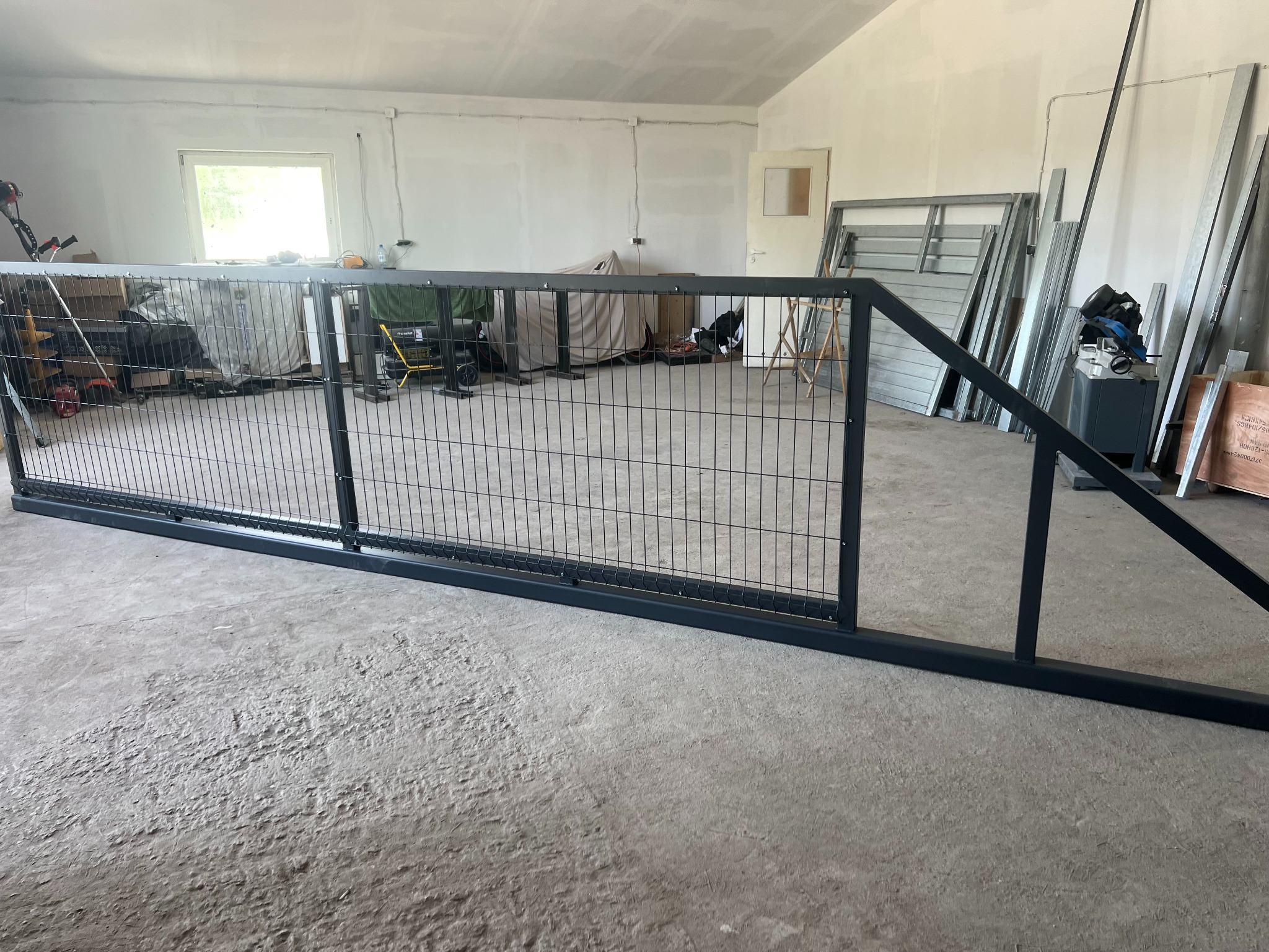 Częściowo zmontowana, przesuwna brama panelowa z czarną ramą w surowym wnętrzu garażu, z elementami konstrukcyjnymi w tle.