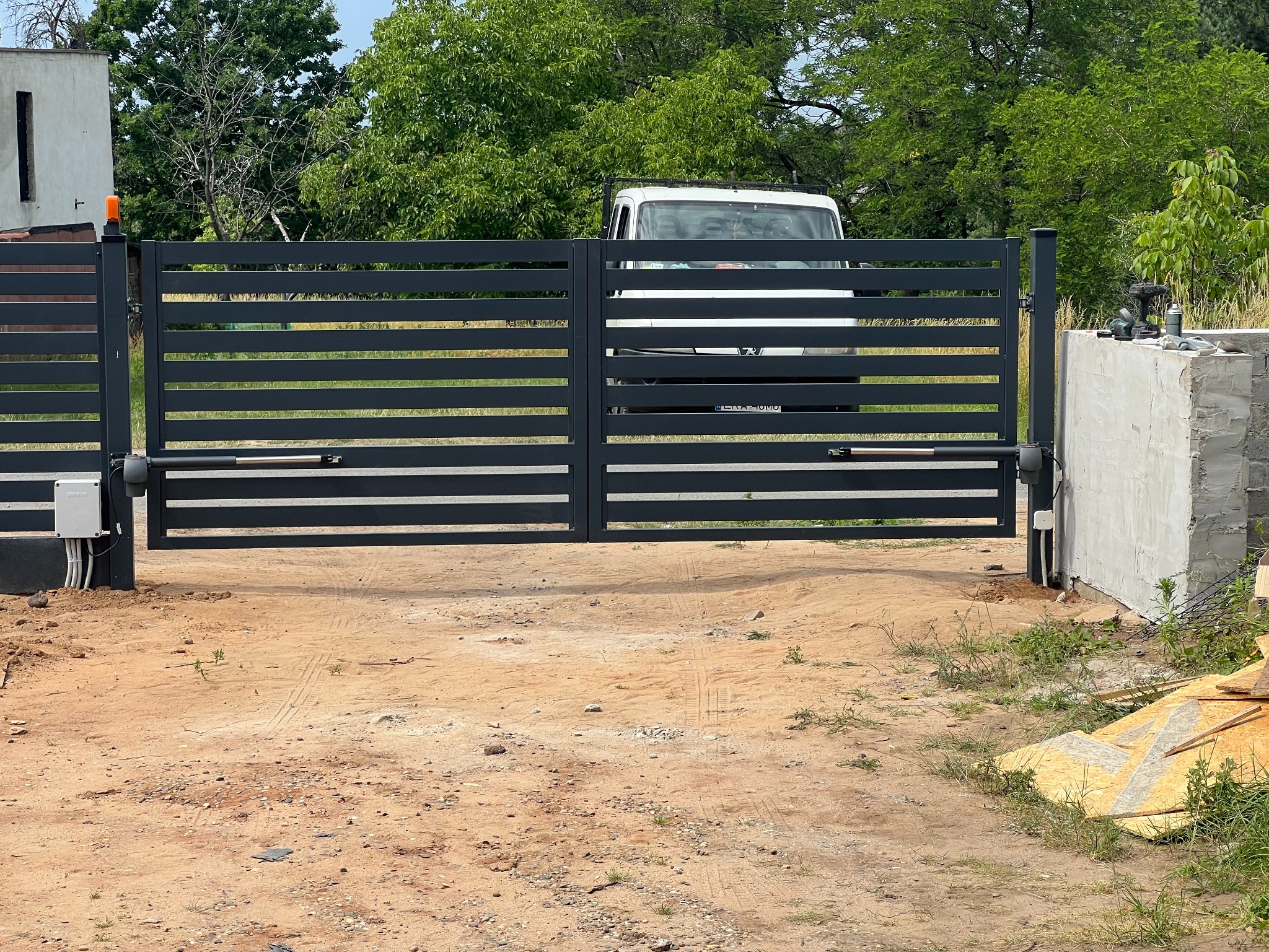 Nowoczesna, pozioma brama wjazdowa z automatyką, widoczna z bliska na tle zieleni i zaparkowanego białego busa. Widoczne narzędzia montażowe.