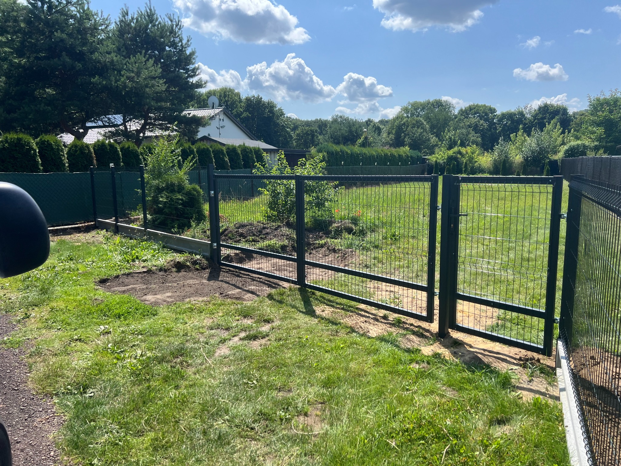 Nowoczesna, grafitowa brama panelowa z furtką, widoczna na tle zielonej trawy i drzew. Element ogrodzenia panelowego z siatki, brama otwarta, słoneczny dzień.