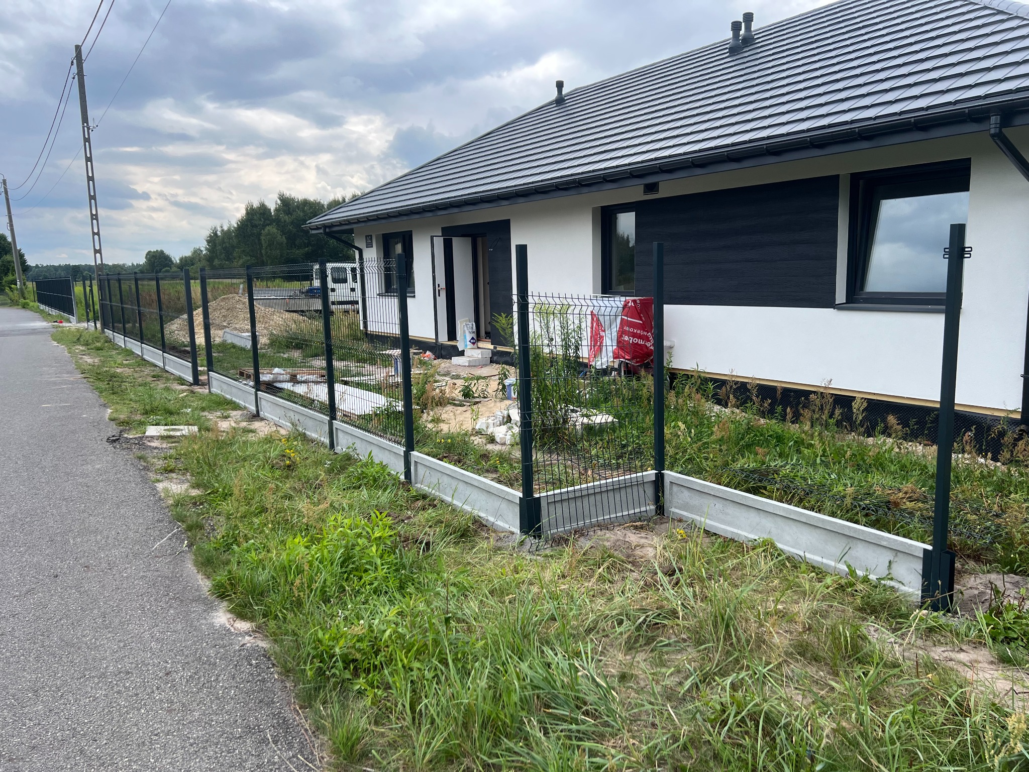 Wznoszenie ogrodzenia panelowego z podmurówką prefabrykowaną przy posesji. Widoczne słupki i częściowo zamontowane panele na tle domu i drogi.