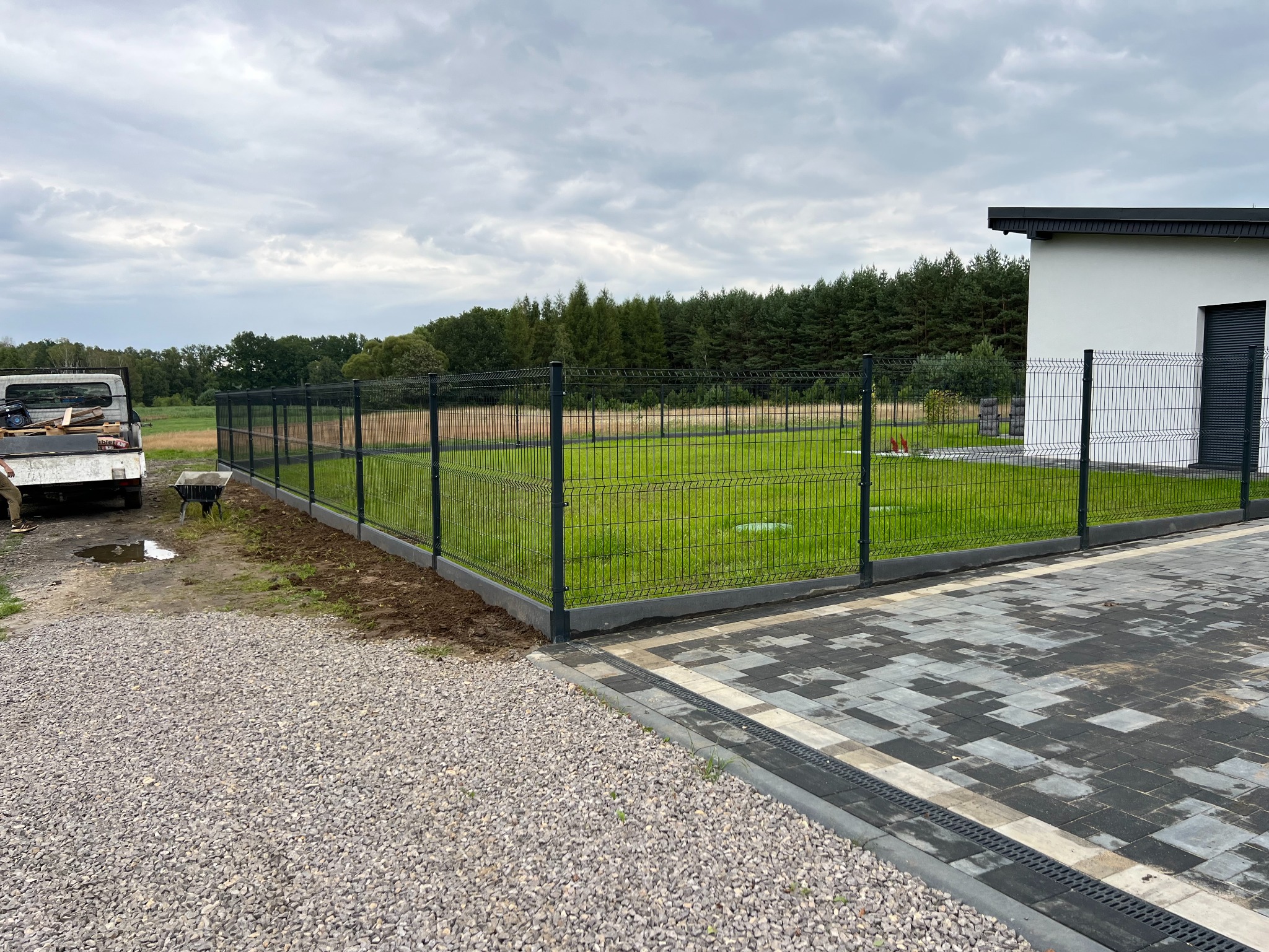 Nowoczesne ogrodzenie panelowe w kolorze grafitowym, oddzielające posesję z trawnikiem od ścieżki z kamienia. Widoczny fragment budynku i samochód dostawczy.