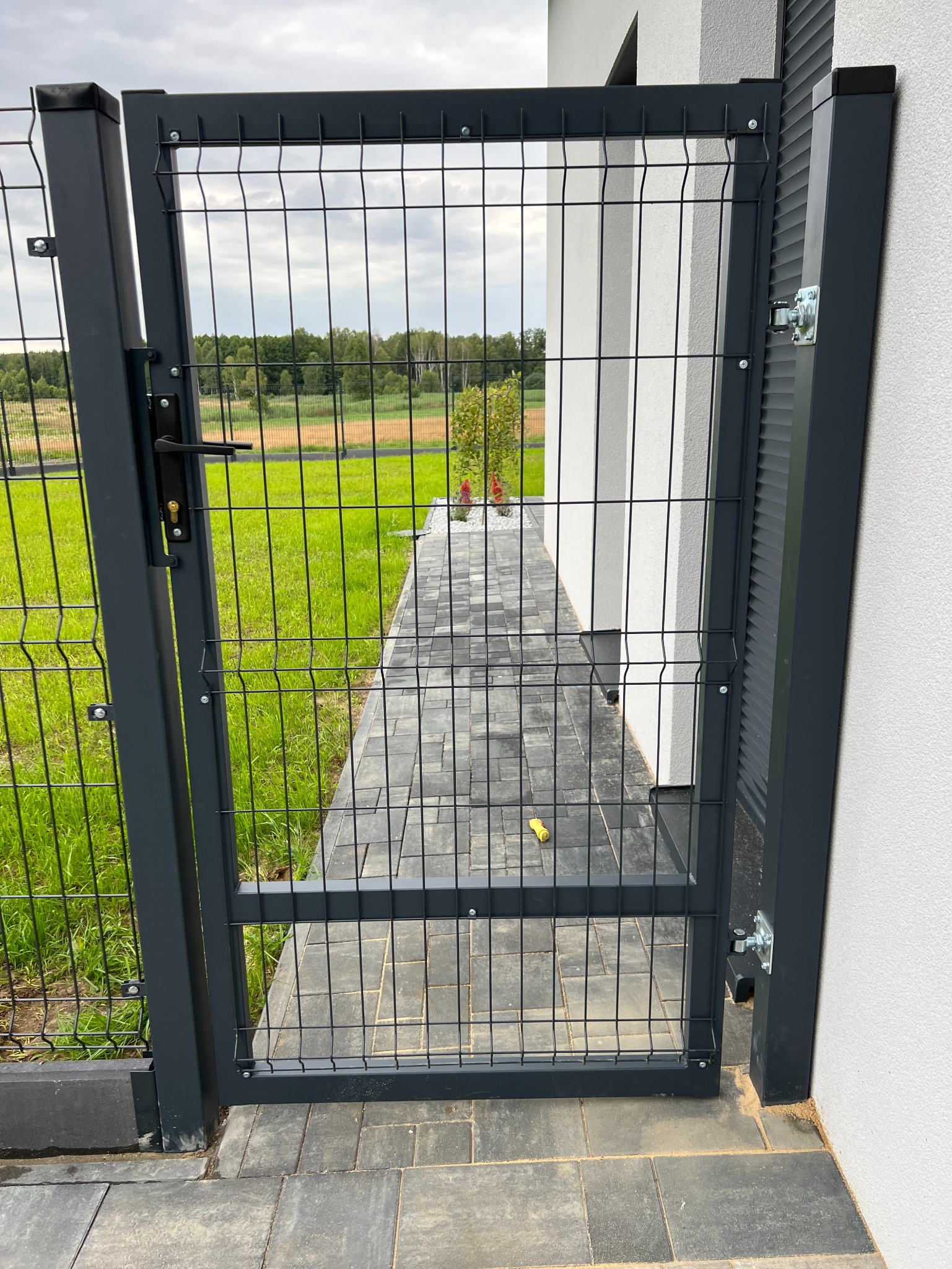 Nowoczesna, grafitowa furtka z siatki panelowej, z widokiem na trawnik i ścieżkę z kostki. Minimalistyczny design, jasna elewacja budynku w tle.