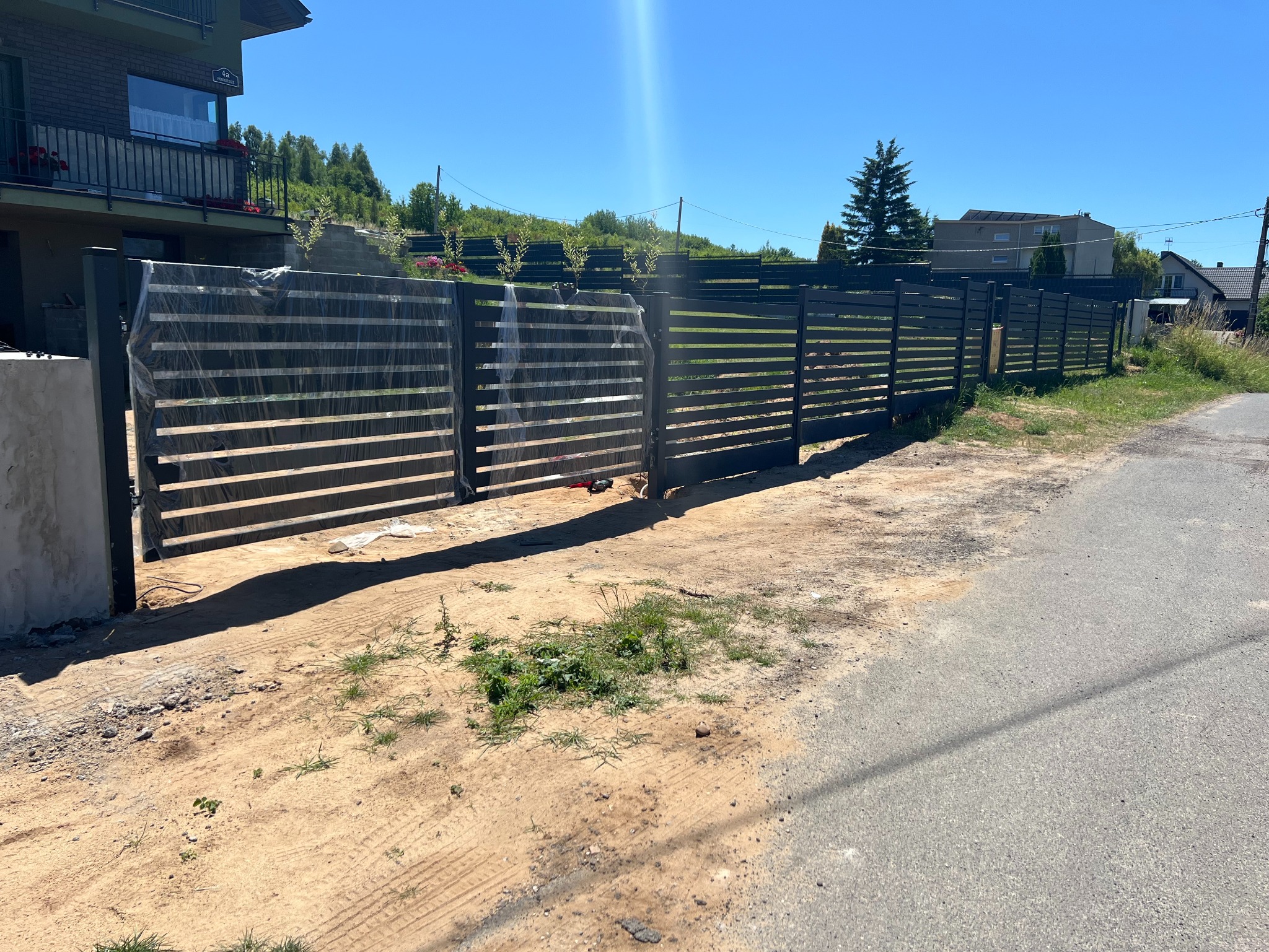 Nowoczesne, poziome ogrodzenie panelowe z czarnych, płaskich elementów, częściowo jeszcze w folii ochronnej, na tle zieleni i błękitnego nieba.