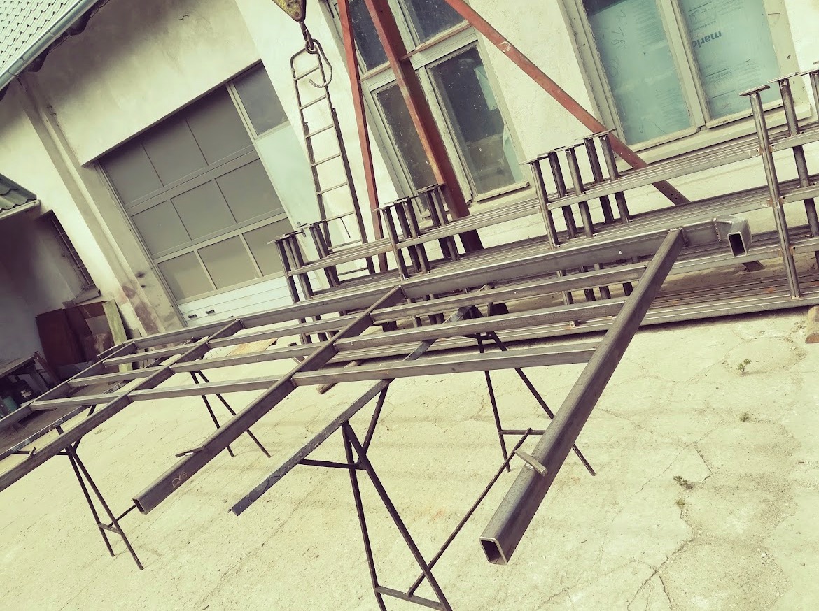 Konstrukcja stalowa z profili kwadratowych, częściowo zmontowana na kozłach, podwieszona na haku dźwigu w warsztacie.