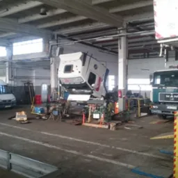 Wnętrze warsztatu samochodowego z podniesioną kabiną ciężarówki na podnośniku, widoczny także MAN oraz inne pojazdy serwisowe.