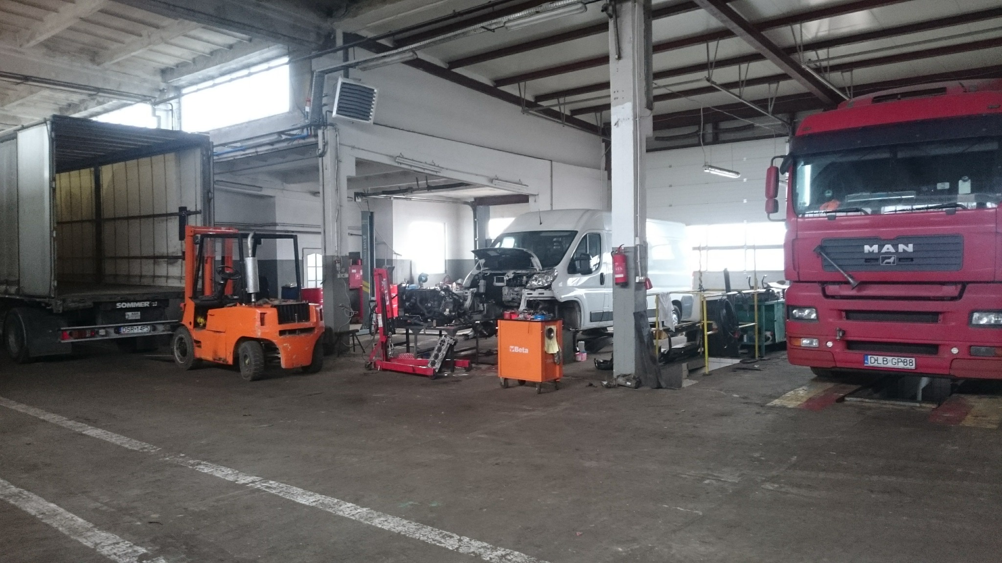 Wnętrze warsztatu samochodowego z widocznym wózkiem widłowym, ciężarówką z naczepą, rozłożonym silnikiem na stojaku, oraz samochodem dostawczym z otwartą maską.