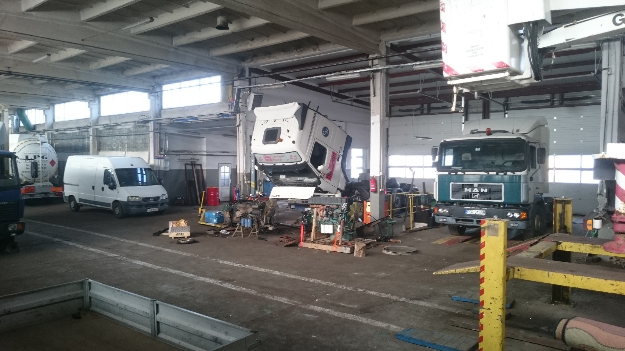 Wnętrze warsztatu samochodowego z podniesioną kabiną ciężarówki na podnośniku, widoczny także MAN oraz inne pojazdy serwisowe.