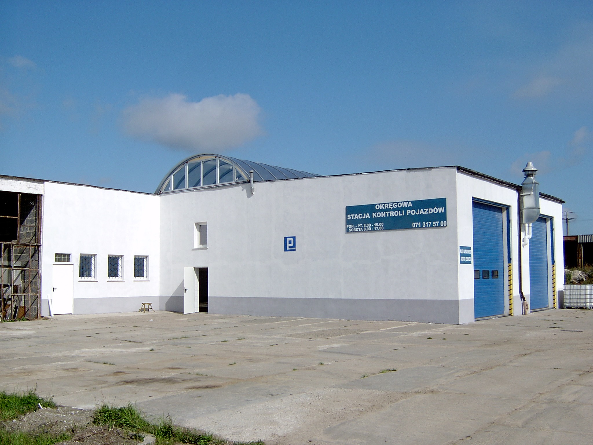 Budynek okręgowej stacji kontroli pojazdów z niebieskimi bramami garażowymi i znakiem parkingowym na białej fasadzie pod błękitnym niebem.