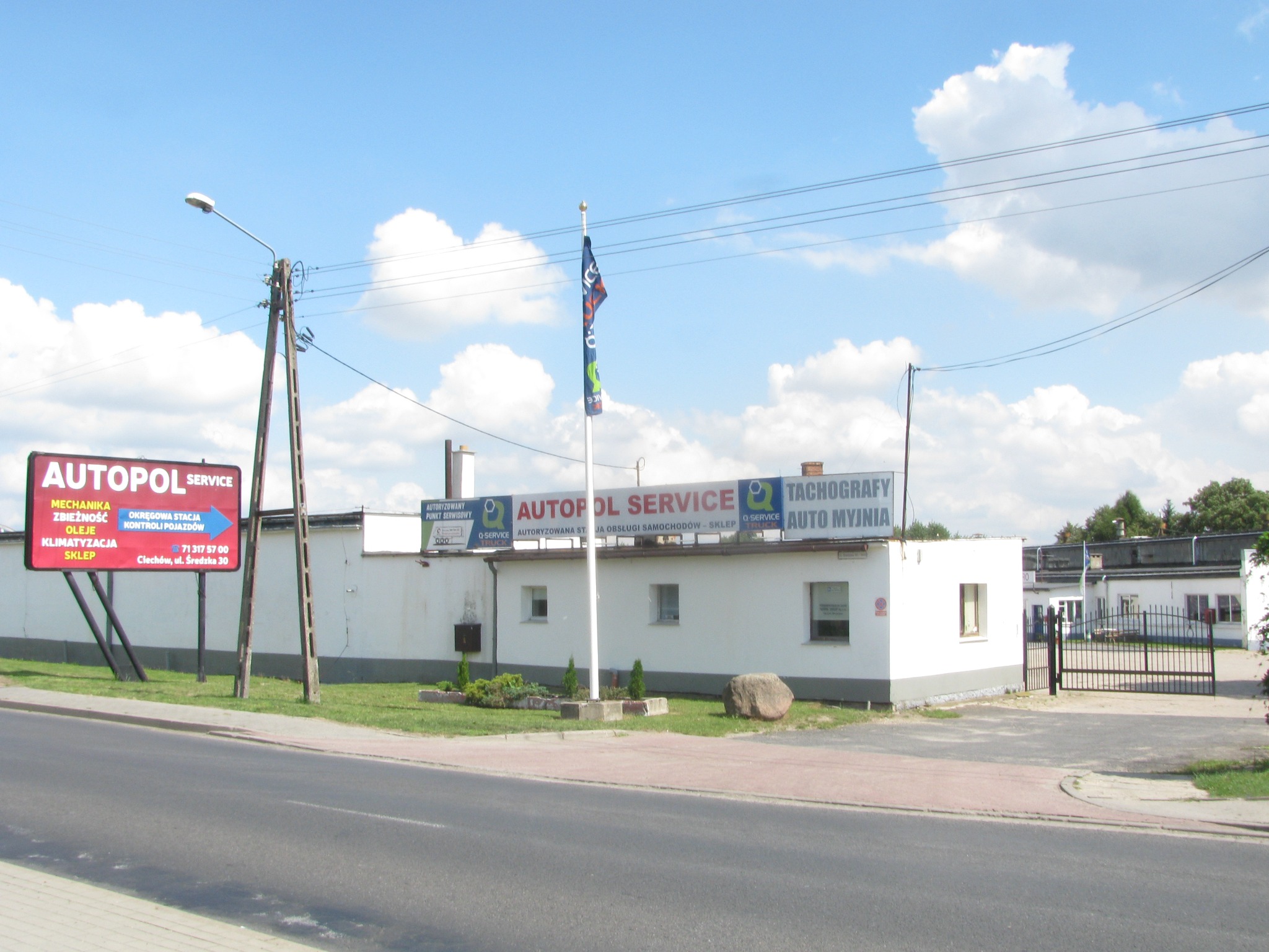 Budynek warsztatu samochodowego Autopol Service z reklamą usług: mechanika, zbieżność, oleje, klimatyzacja, sklep, okręgowa stacja kontroli pojazdów. Na dachu baner z napisem Autopol Service...