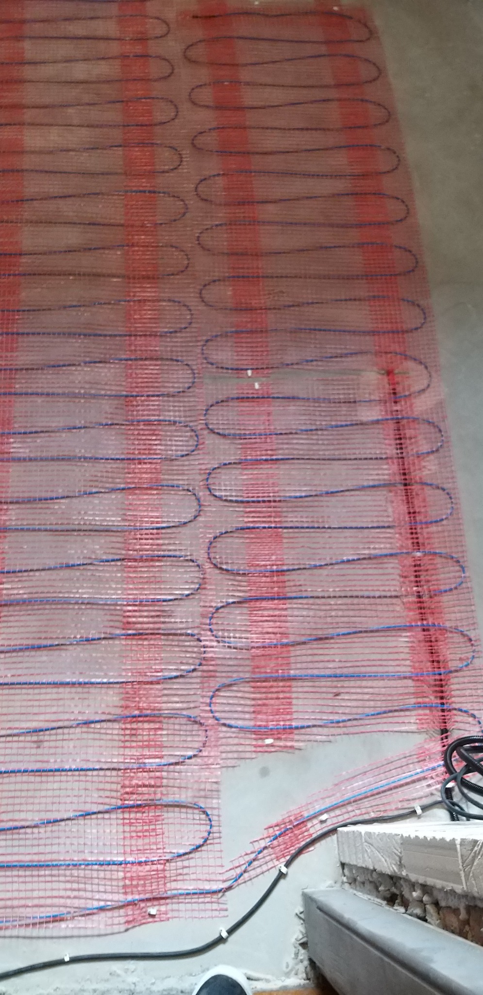 Rozłożona mata grzewcza z niebieskim przewodem na czerwonej siatce, widoczne kable zasilające i fragment posadzki.