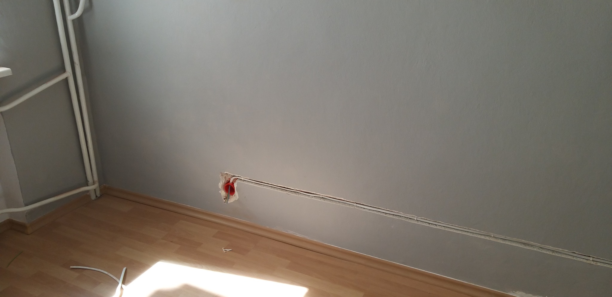 Szara ściana z widocznymi przewodami elektrycznymi w listwach i puszką instalacyjną bez osprzętu, jasna podłoga z paneli, rury grzejnika przy ścianie.