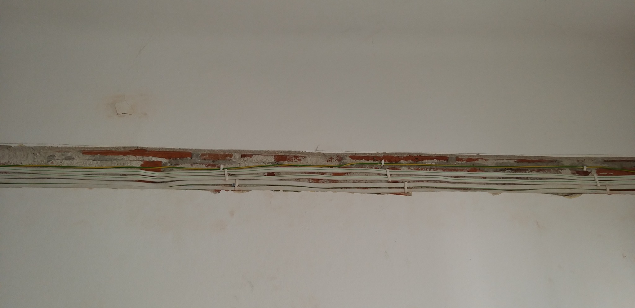 Białe kable elektryczne ułożone w bruździe w ścianie z czerwonej cegły, widoczne przewody żółto-zielone, ściana pomalowana na biało, widoczne ślady po szpachlowaniu.