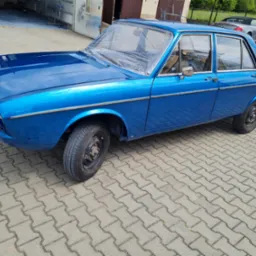 Błękitny, odrestaurowany samochód marki Audi 100, z folią ochronną na przedniej szybie, zaparkowany na kostce brukowej przed garażem. W tle widoczny inny pojazd.