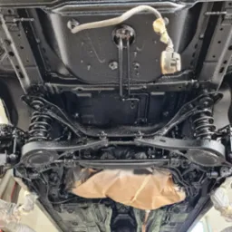 Podwozie samochodu po zabezpieczeniu antykorozyjnym, widoczne elementy zawieszenia i układu wydechowego, niekt&oacute;re fragmenty osłonięte papierem maskującym, świeżo pomalowane na czarno, widok od dołu.