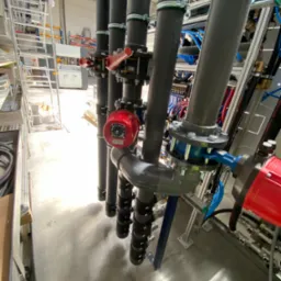 System rur PVC-U d110 z czerwonymi zaworami i niebieskimi elementami, widoczne zejście do maszyny w hali produkcyjnej.