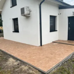 Nowo ułożona, brązowa kostka brukowa przed białym budynkiem z czarnymi oknami i drzwiami, widoczne obrzeża i fragment terenu z ziemią.
