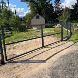 Nowoczesna, metalowa brama panelowa z siatki, pomalowana na ciemny kolor, zamontowana na betonowej podmur&oacute;wce, z widocznym zamkiem. W tle zielony trawnik, drewniany domek letniskowy z basenem...