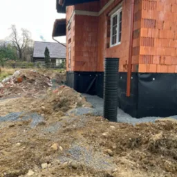 Surowy stan budowy domu z czerwonej cegły: widoczne fundamenty zabezpieczone czarną folią izolacyjną i pionowa rura drenażowa, teren wokół z gruzem i ziemią.