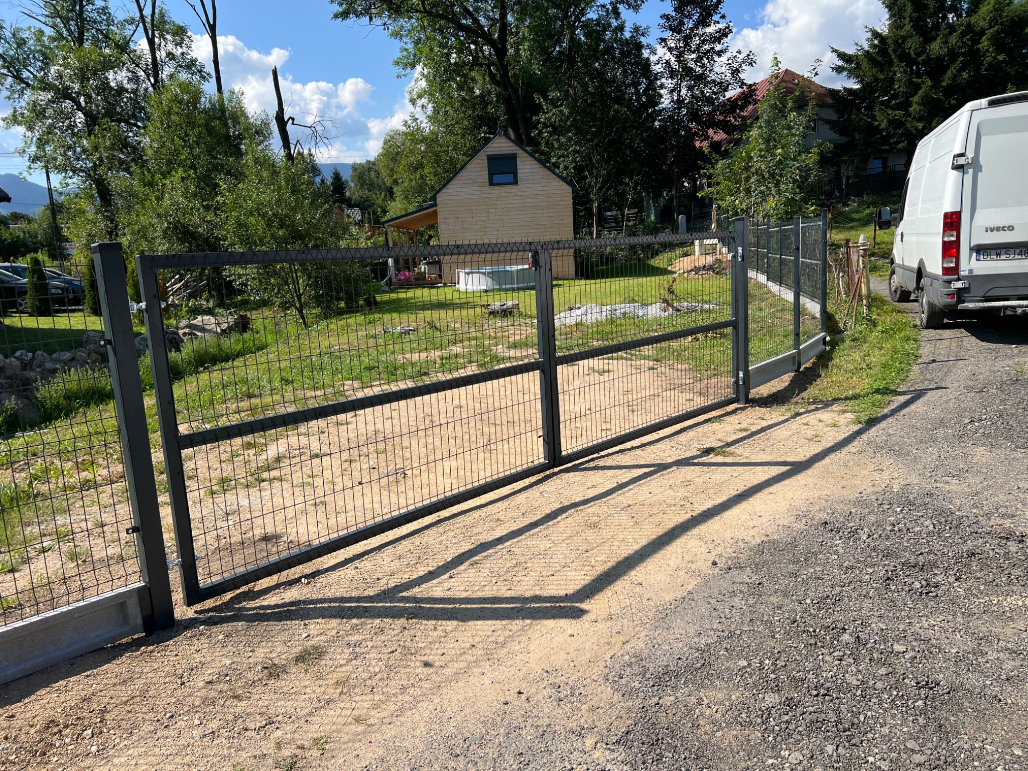 Nowoczesna, metalowa brama panelowa z siatki, pomalowana na ciemny kolor, zamontowana na betonowej podmurówce, z widocznym zamkiem. W tle zielony trawnik, drewniany domek letniskowy z basenem...
