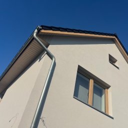 R.S - Budownictwo - Fragment elewacji domu jednorodzinnego z jasnym tynkiem, widoczne okna z drewnianą stolarką, rynna spustowa i fragment dachu z czarną dachówką na tle błękitnego nieba.
