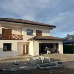 Dwukondygnacyjny dom jednorodzinny z białymi ścianami elewacyjnymi, brązowymi oknami i balkonem z drewnianą balustradą; na podjeździe leżą elementy rusztowania, a w otwartym garażu widać pracownika...