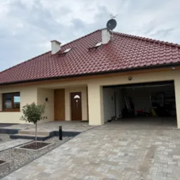 Elewacja domu jednorodzinnego w beżowym kolorze z czerwoną dachówką, widok na podjazd z kostki brukowej i otwarty garaż. Nowoczesny styl.