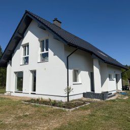 R.S - Budownictwo - Nowoczesny dom jednorodzinny z białą elewacją i ciemną dachówką, widok z ogrodu w słoneczny dzień. Mały ogródek przed domem.