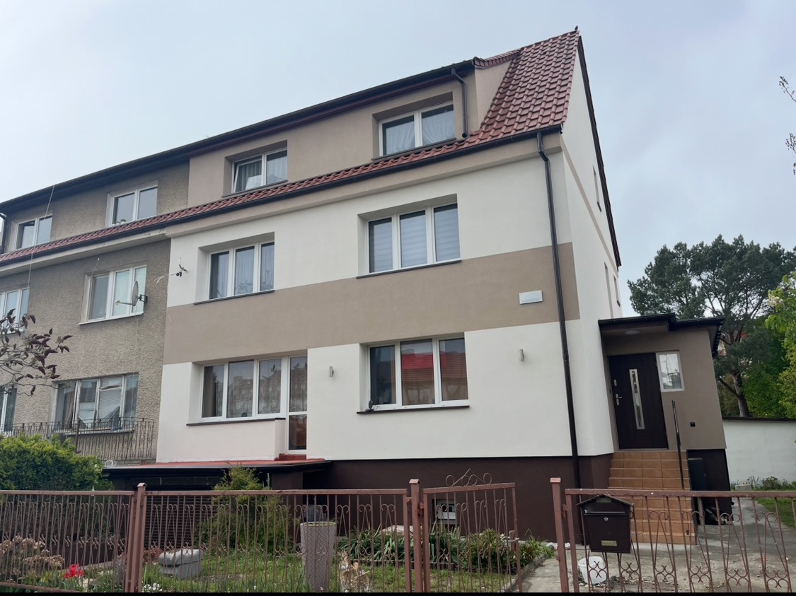 Częściowo odnowiony budynek wielorodzinny z widoczną różnicą w wyglądzie elewacji przed i po renowacji, z brązowym pasem na elewacji i czerwoną dachówką.