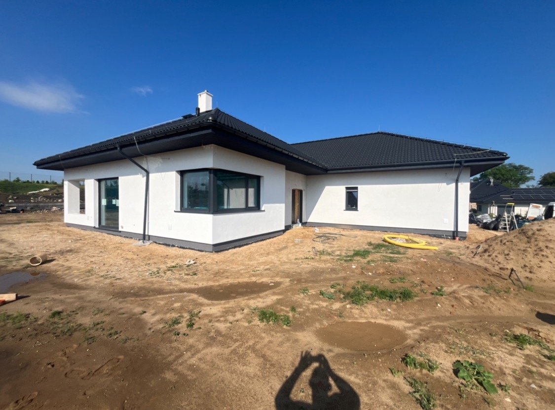 Nowoczesny dom jednorodzinny z białą elewacją i czarnym dachem, widoczne niedokończone prace ziemne wokół budynku oraz żółty wąż na trawniku.