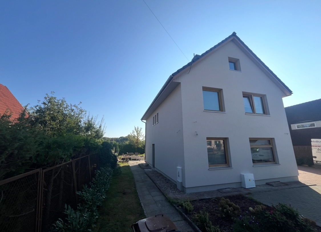 Dwu kondygnacyjny dom z jasną elewacją po renowacji, z widocznymi oknami z drewnianymi ramami i skrzynką elektryczną przy ścianie, otoczony zielenią i niskim płotem.