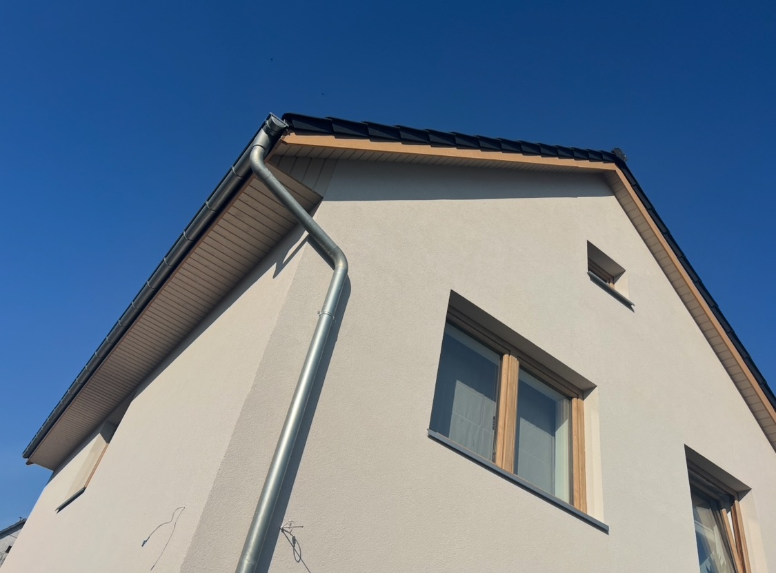 Fragment elewacji domu jednorodzinnego z jasnym tynkiem, widoczne okna z drewnianą stolarką, rynna spustowa i fragment dachu z czarną dachówką na tle błękitnego nieba.