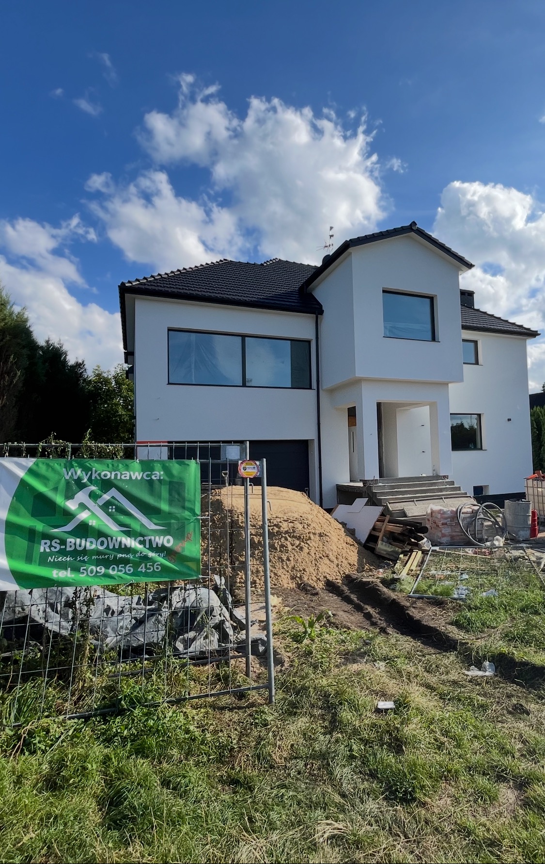 Nowoczesny, biały dom jednorodzinny w Dobra, z widocznym stosem piasku i ogrodzeniem budowy, okna zabezpieczone folią, elewacja w trakcie realizacji.