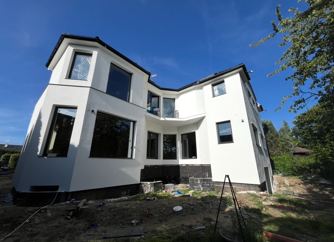 Nowoczesny, biały dom z czarnymi ramami okiennymi, po ociepleniu i tynkowaniu elewacji, widok z dołu na tle błękitnego nieba, teren wokół domu w trakcie prac wykończeniowych.