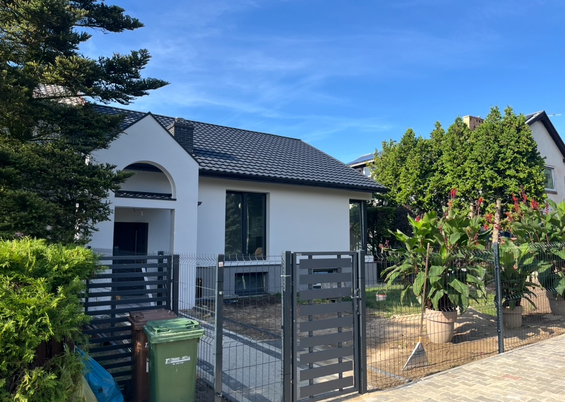 Nowoczesny dom jednorodzinny z białymi ścianami i grafitowym dachem, widoczny zza ogrodzenia z elementami poziomymi, z dwoma koszami na śmieci po lewej stronie bramki i roślinami w donicach po prawej.
