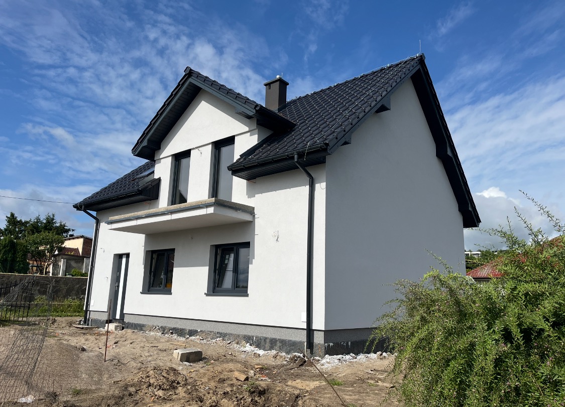 Nowoczesny, dwukondygnacyjny dom z szarą elewacją i czarną stolarką okienną, widok z zewnątrz na tle błękitnego nieba z chmurami, teren wokół domu w trakcie prac wykończeniowych.